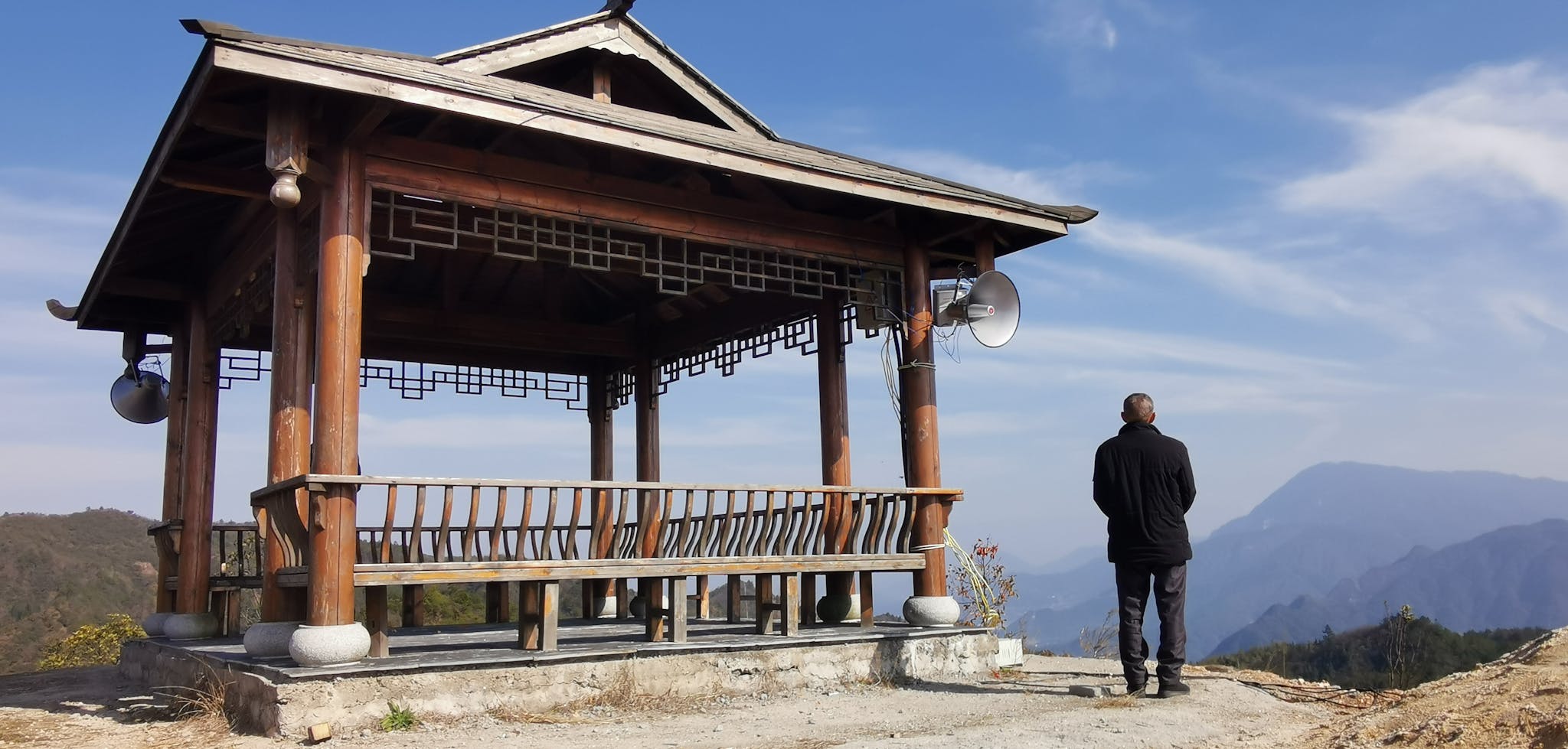 A lone figure stands by a traditional Chinese pavilion on a mountain on a clear day.